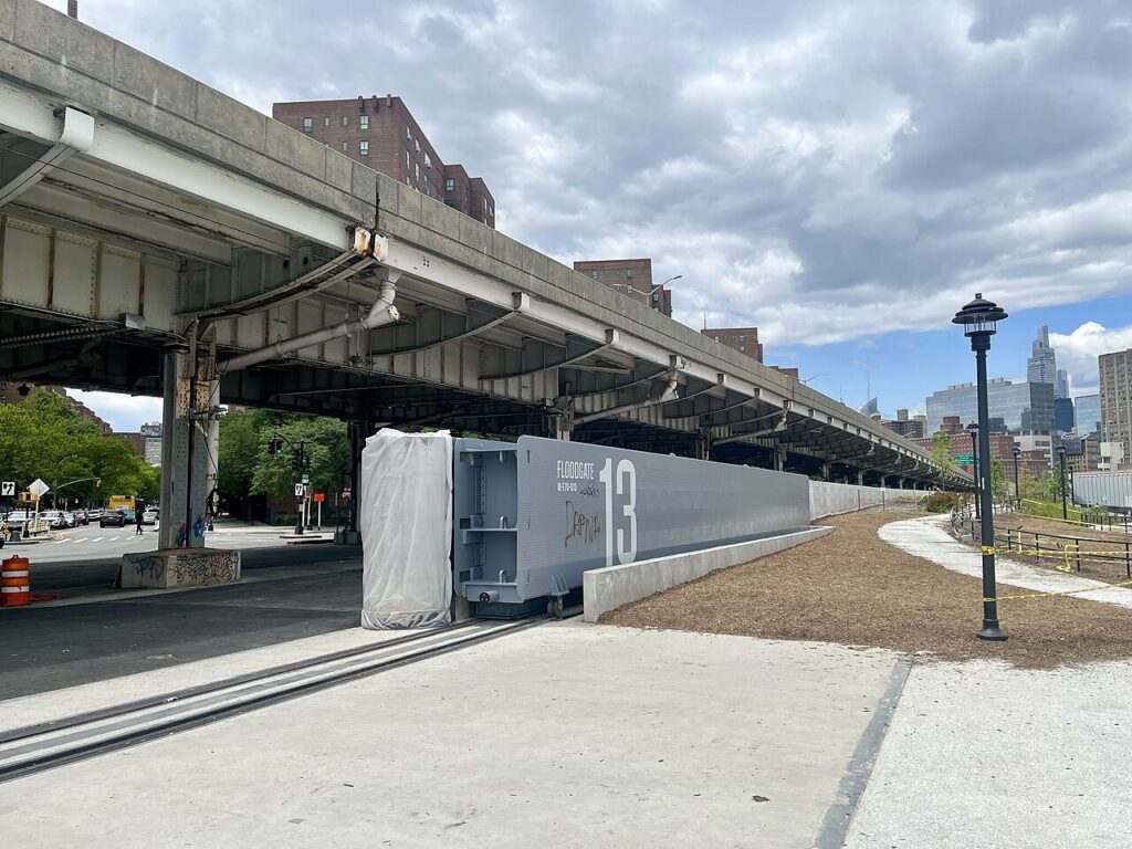 A large sliding floodgate beside tracks crossing a paved entrance beneath an elevated roadway.